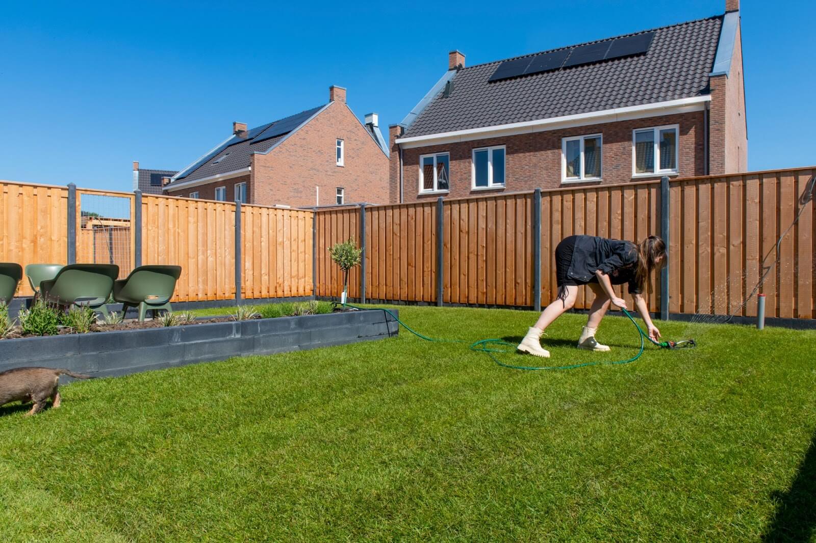 A woman is watering her grass in a backyard that has brown fencing.