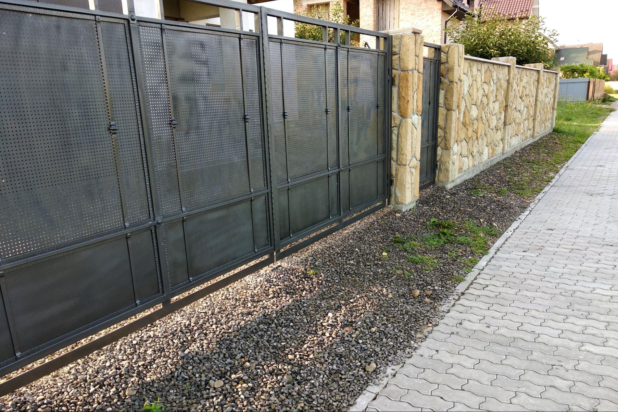 A custom natural stone fence with a black metal gate bordered by cement slab pavement.