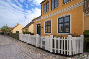 A yellow and white house with elegant architecture, enclosed by classic white wood fencing materials.
